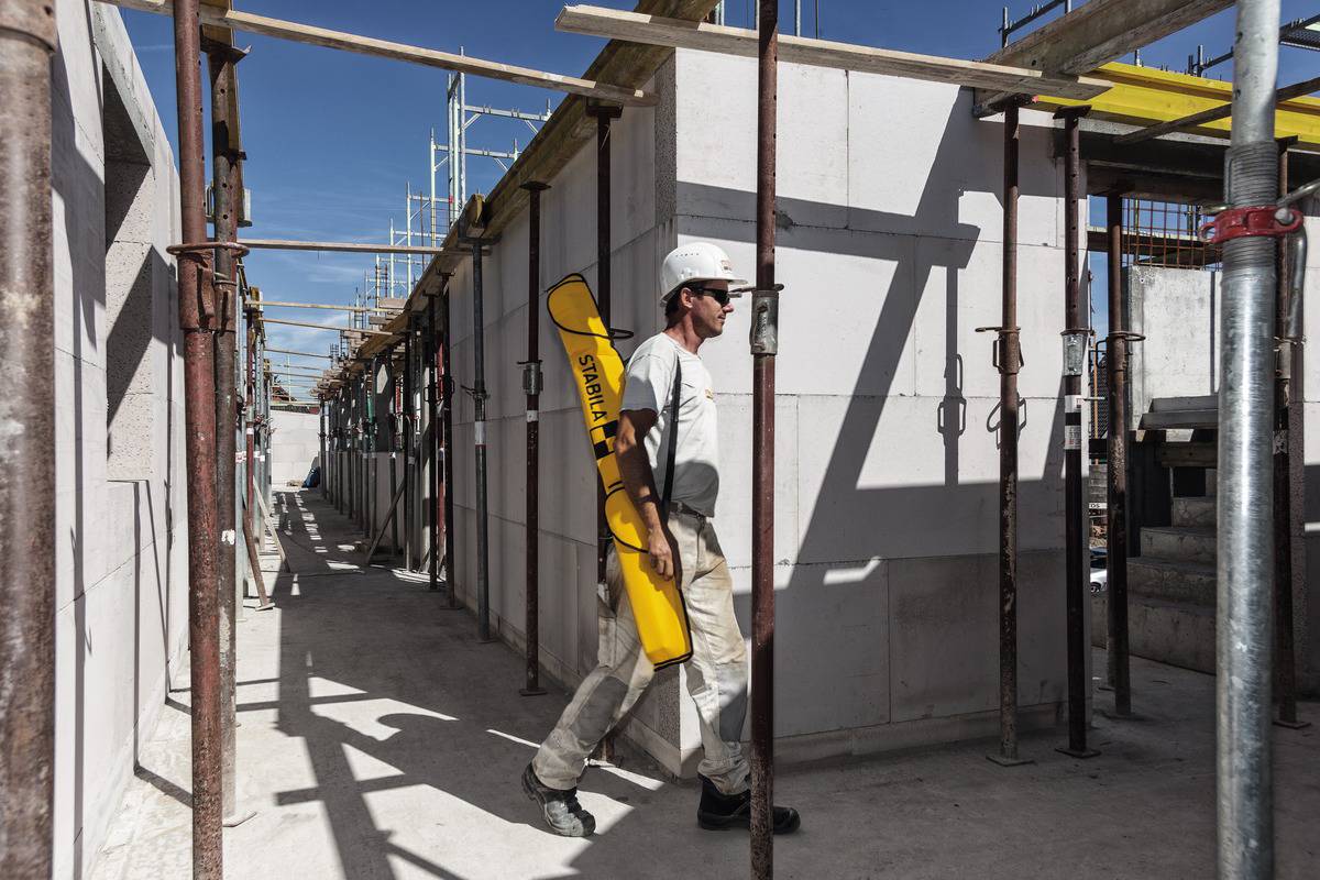 Un ouvrier en tenue de protection porte un sac à outils jaune sur un chantier de construction avec des échafaudages et des murs en cours de construction sous un ciel bleu.