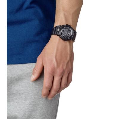 La personne porte une montre-bracelet noire et robuste au poignet gauche. Un t-shirt bleu et un pantalon gris sont visibles en bas.