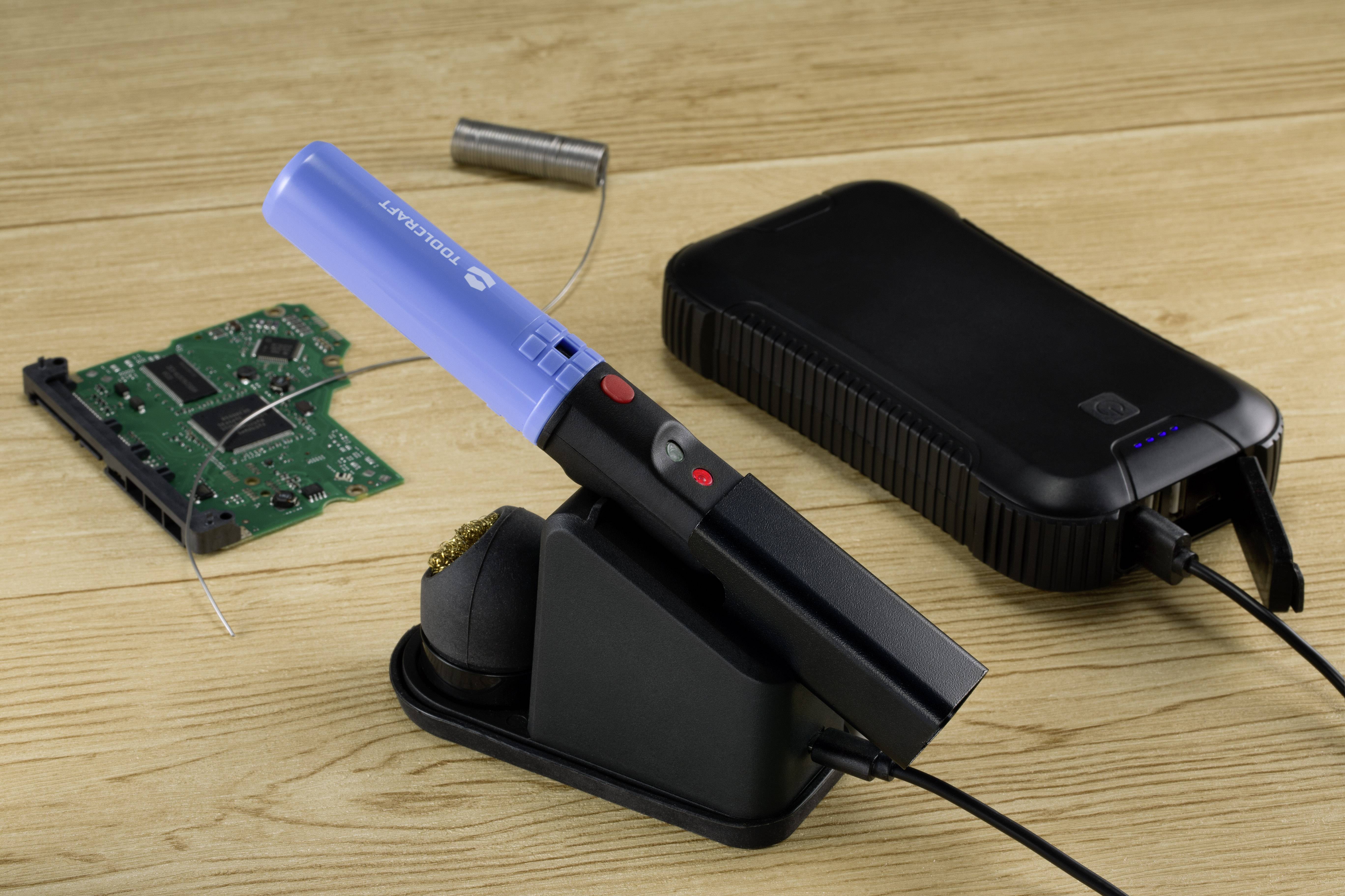 Fer à souder à pointe bleue, branché à un appareil noir, à côté d'une carte électronique verte et d'accessoires de soudure sur une table en bois.