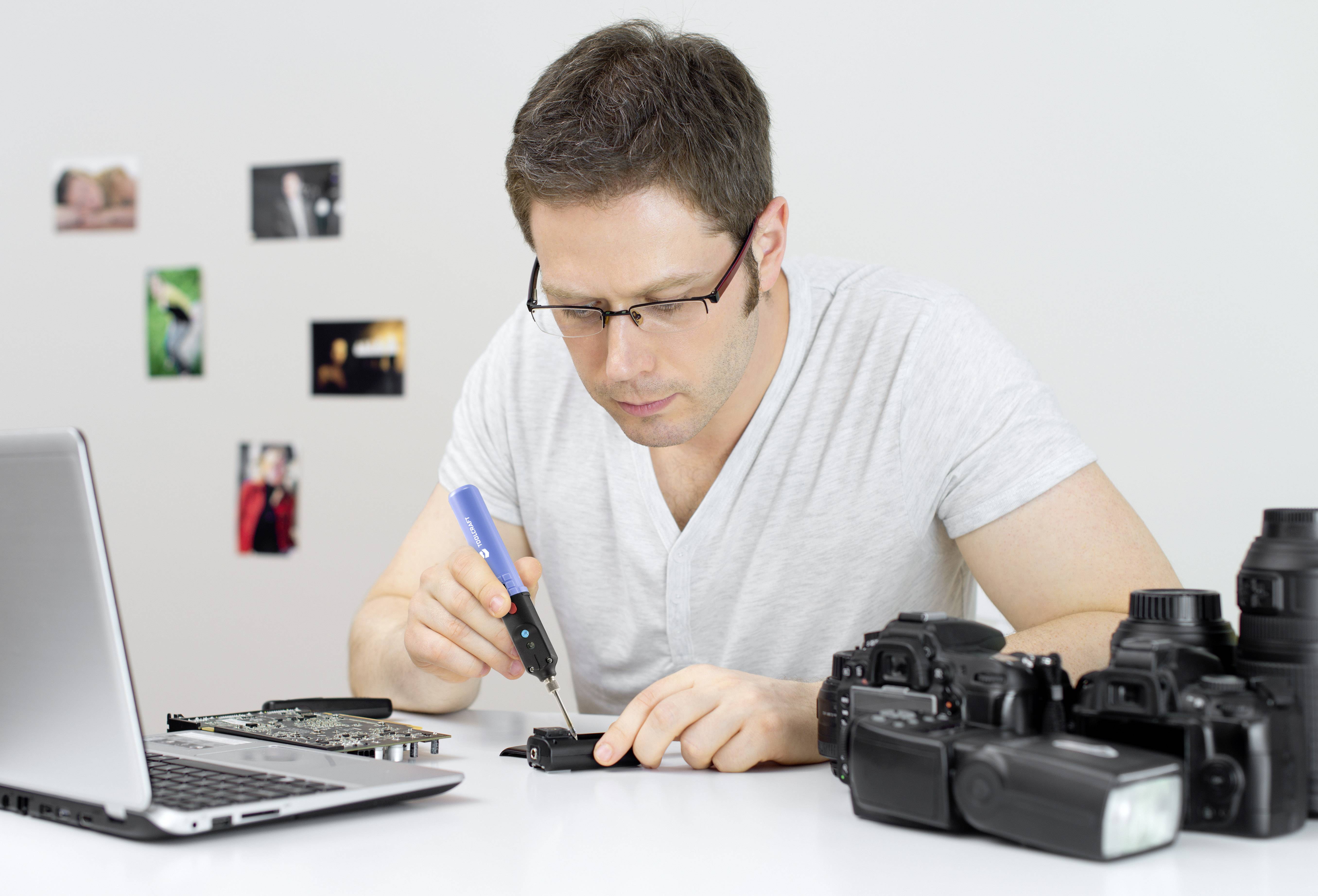 'Un homme répare un appareil photo avec un fer à souder sur un bureau. Un ordinateur portable et des accessoires photo sont posés sur la table. Des photos sont accrochées au mur en arrière-plan.'