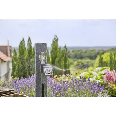 Un système d'irrigation avec minuterie devant un jardin de lavande et d'autres plantes. Des arbres sont visibles en arrière-plan.