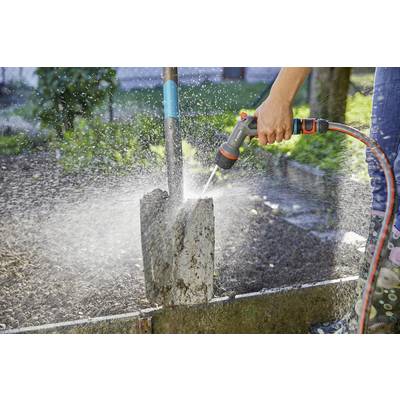 Une personne nettoie une pelle sale avec un tuyau d'arrosage dans un jardin. Des gouttes d'eau éclaboussent dans les airs.