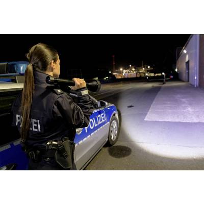 Une policière se tient la nuit à côté d'un véhicule de service et éclaire une zone sombre avec une lampe de poche.