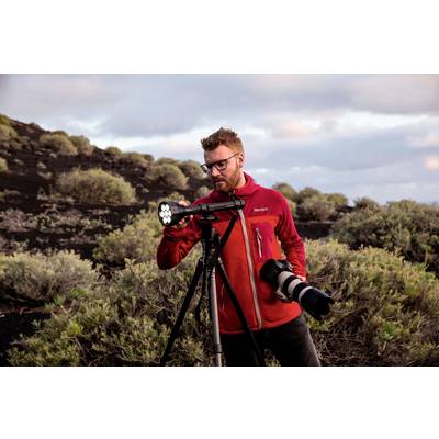 Un homme portant une veste rouge se tient avec un appareil photo sur un trépied devant un paysage de buissons et de terre sombre sous un ciel nuageux.
