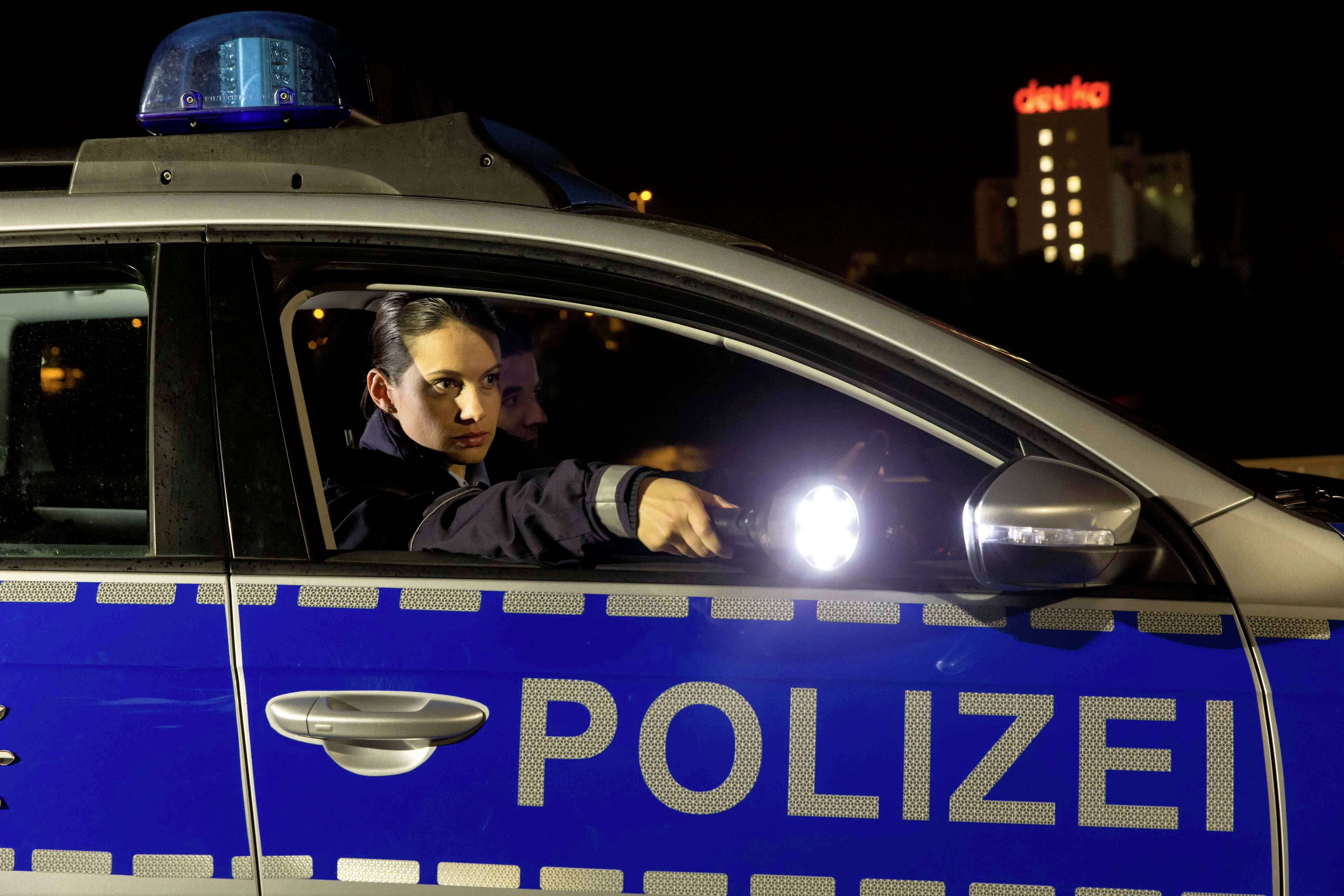 Une policière est assise dans une voiture de patrouille et braque une lampe de poche. Un bâtiment éclairé est visible en arrière-plan.