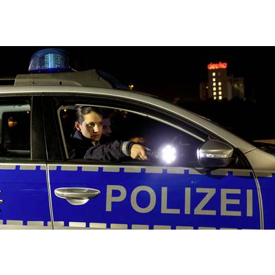 Une policière est assise dans une voiture de patrouille et braque une lampe de poche. Un bâtiment éclairé est visible en arrière-plan.