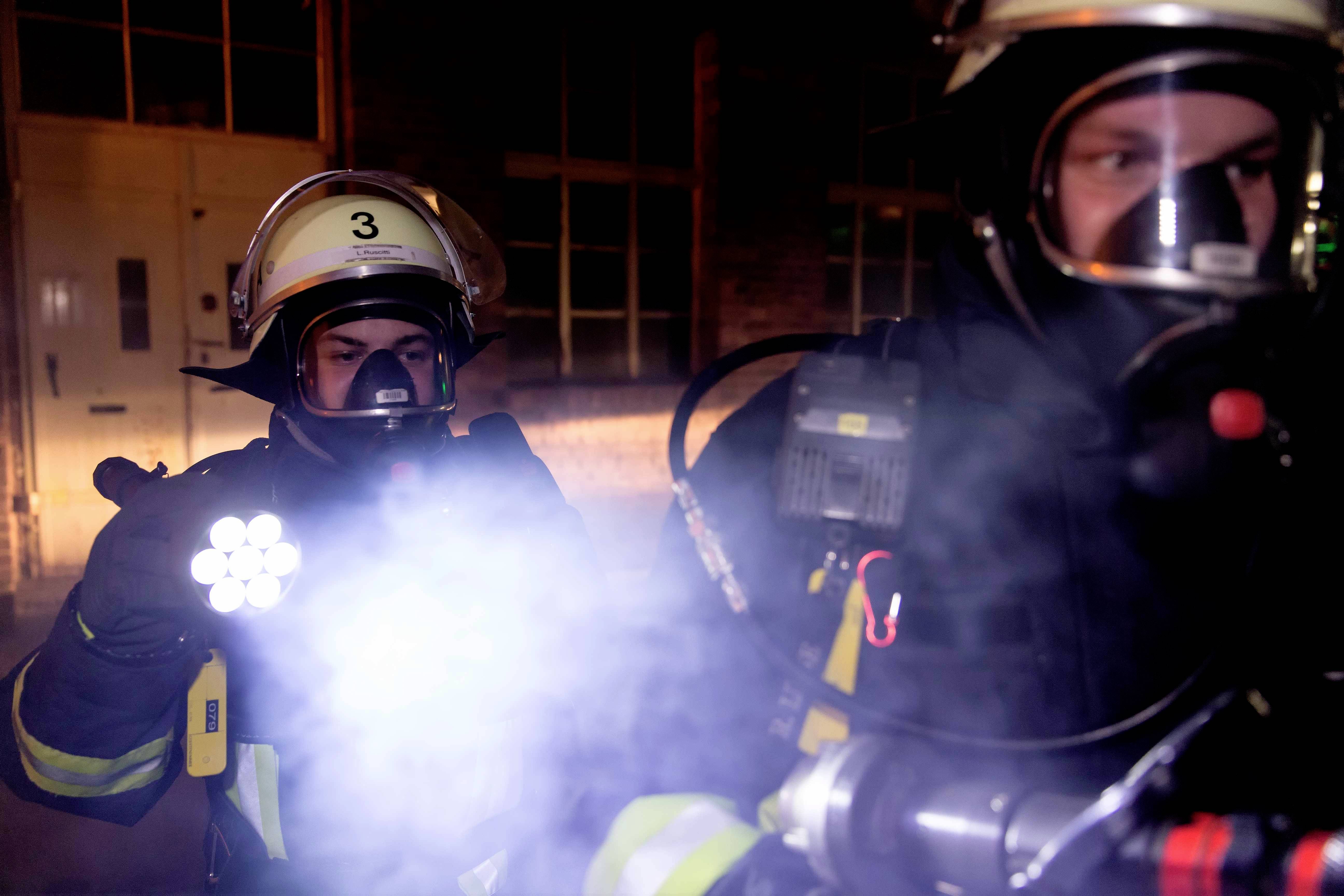 Deux pompiers entièrement équipés en intervention, l'un tenant une lampe torche brillante devant lui, entourés de fumée.