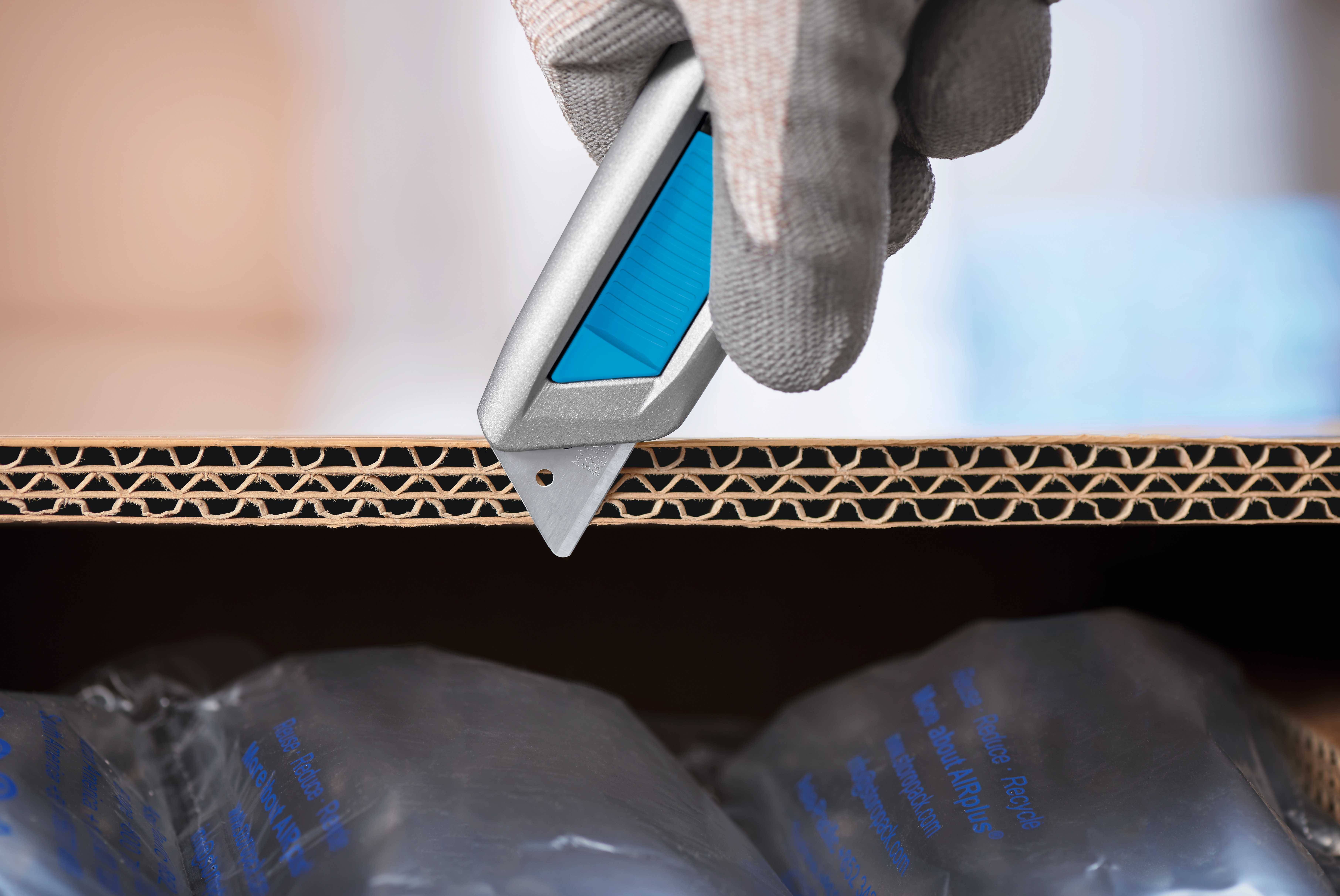 Une main gantée tient un cutter qui entaille un carton ondulé. En dessous se trouvent des emballages à bulles d'air.