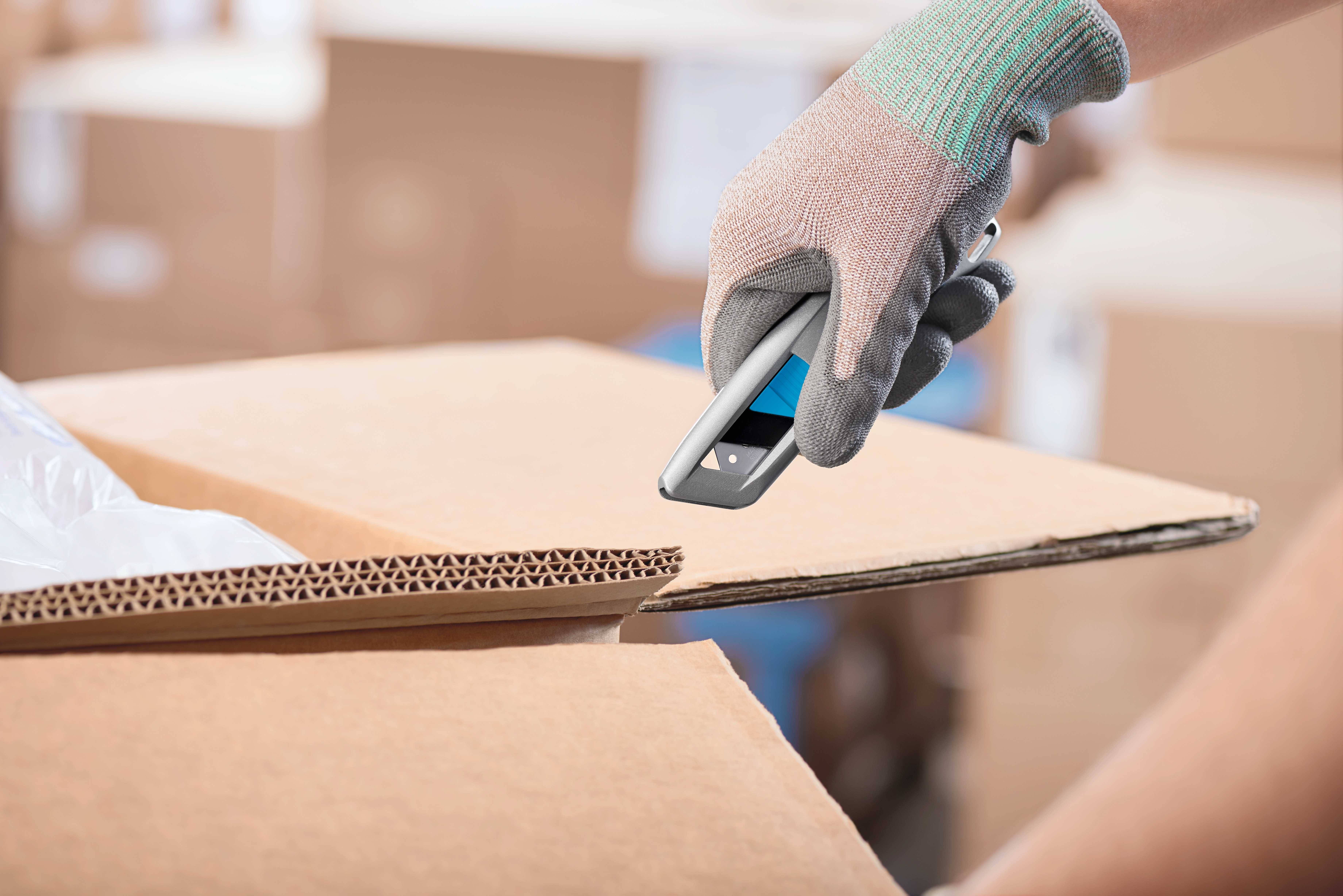 Une personne portant des gants ouvre prudemment un carton à l'aide d'un cutter dans un environnement d'entrepôt.