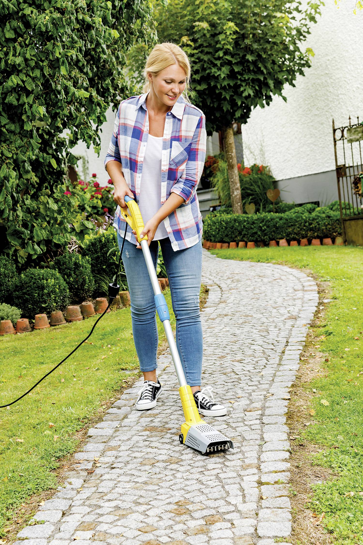 Une femme utilise un appareil électrique pour éliminer les mauvaises herbes sur un chemin dallé de jardin. Des arbres et une pelouse sont visibles en arrière-plan.