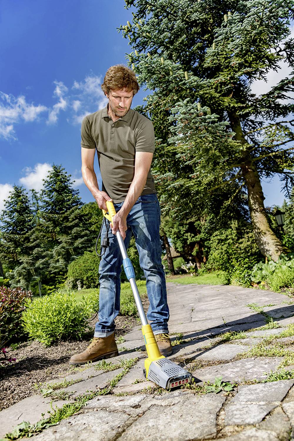Une personne nettoie un chemin pavé dans un jardin à l'aide d'un appareil électrique. Des arbres et des plantes sont visibles en arrière-plan.