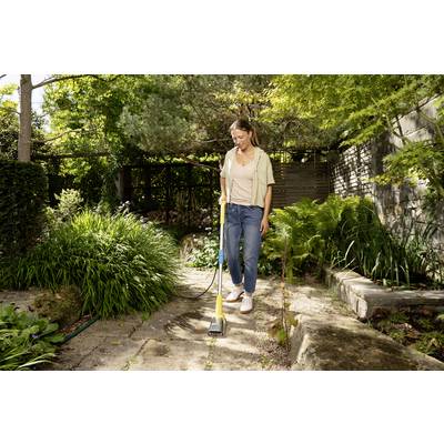 Femme balayant un chemin de jardin en pierre avec un balai dans une cour arrière feuillue.