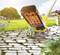 Une brosse désherbeuse enlève les mauvaises herbes entre les pavés dans un jardin ensoleillé. En arrière-plan, on aperçoit des parterres de fleurs et des arbres.