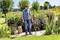 Un homme portant une chemise bleue et un jean utilise un désherbant sur un chemin de jardin. Des fleurs et des arbres en fleurs sont visibles en arrière-plan.