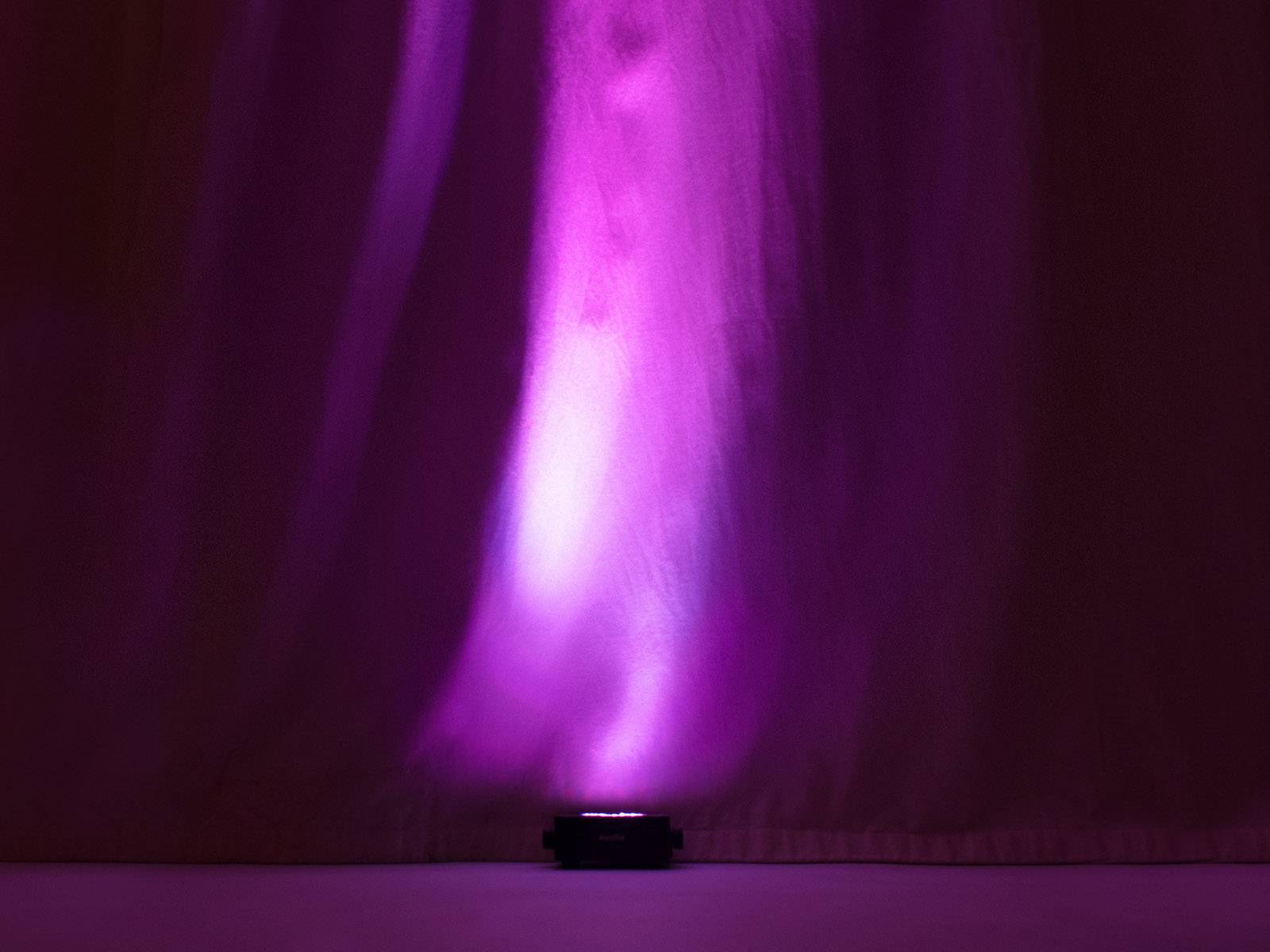 Un projecteur projette une lumière violet-rose sur un rideau, créant des ombres douces et des dégradés de couleur.