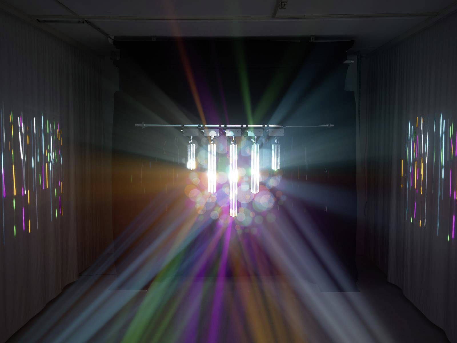 Installation lumineuse dans une pièce sombre, des rayons colorés et des réflexions se propagent à partir de sources lumineuses centrales vers l'extérieur.