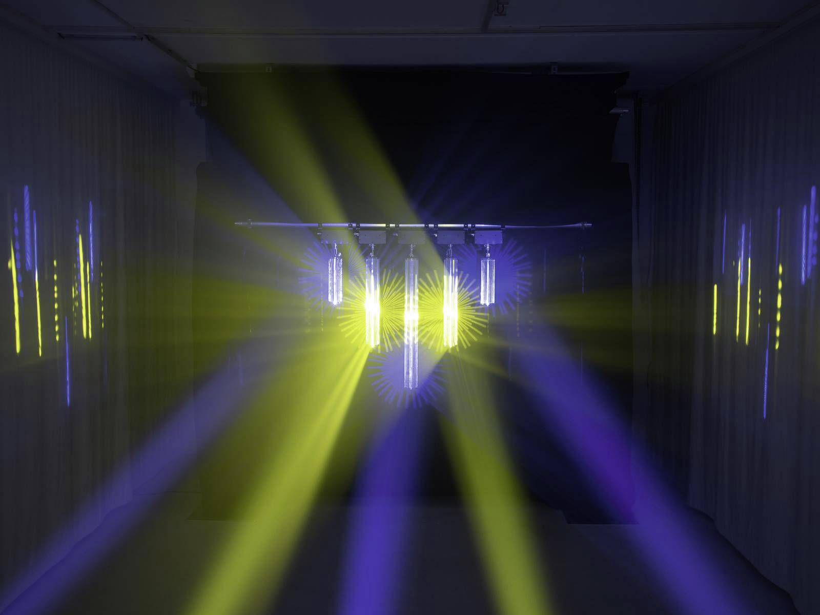 Installation lumineuse dans une pièce sombre avec des rayons jaunes et bleus émanant de plusieurs tubes, illuminant les murs.
