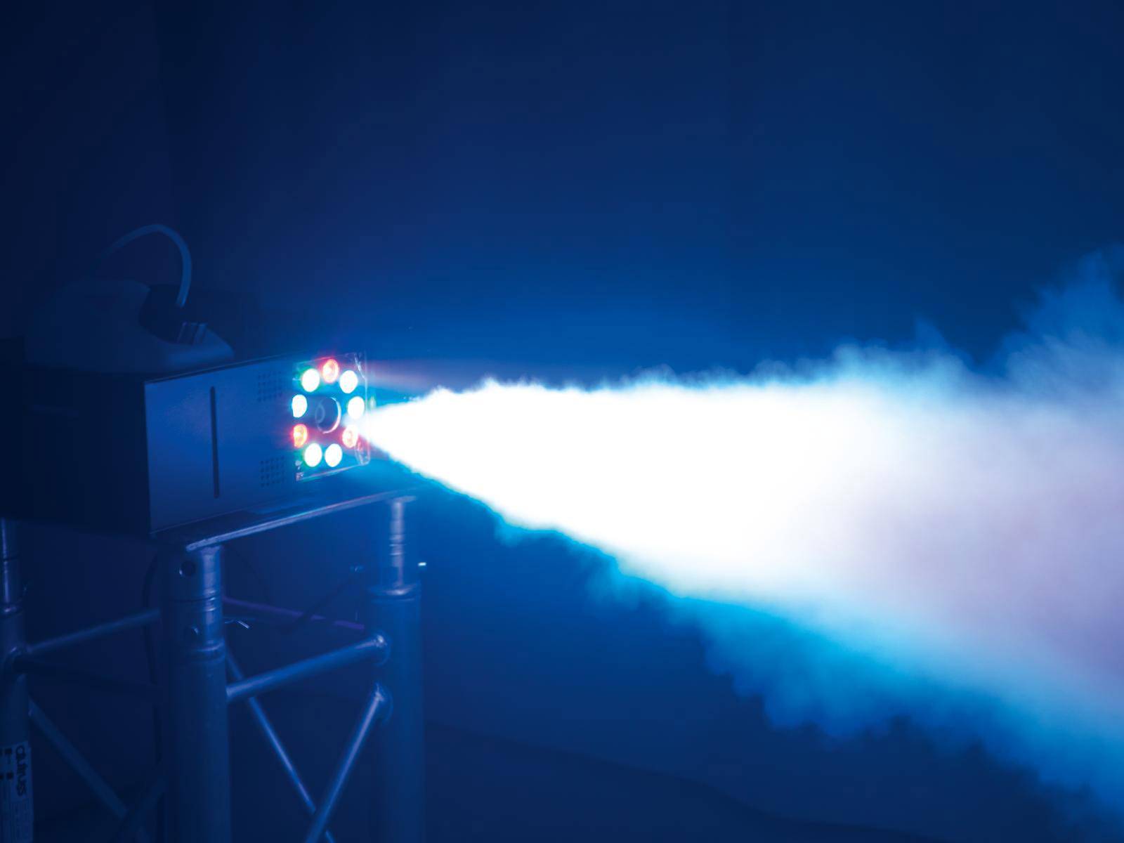 Machine à fumée produit un faisceau lumineux brillant dans un environnement sombre, montée sur un trépied métallique. Un cône de lumière et de brume sont visibles.