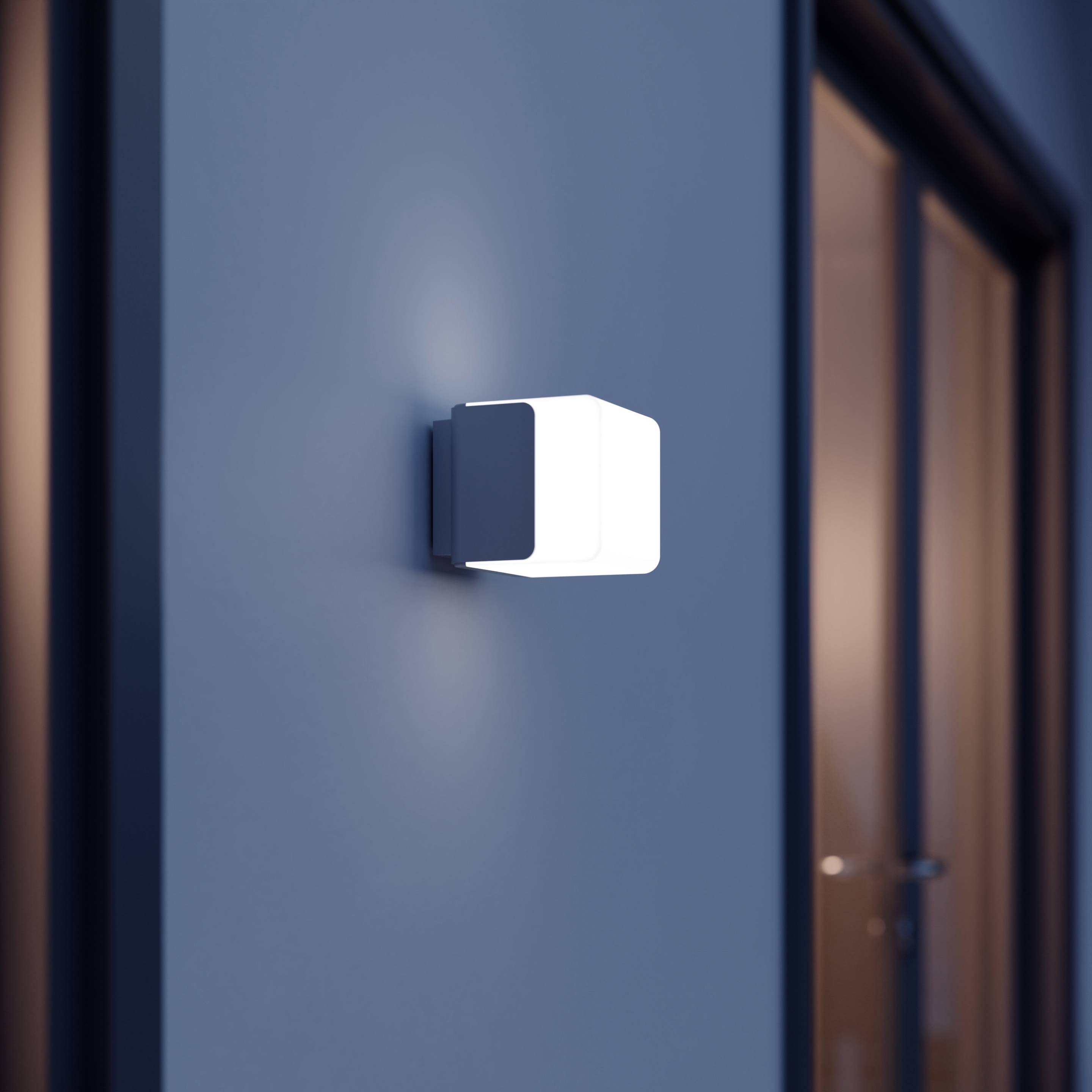 Un luminaire mural moderne est fixé sur un mur extérieur gris foncé. Il présente un design épuré et carré, diffusant une lumière douce.