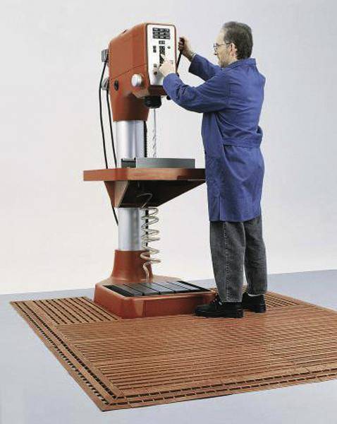 Un homme en blouse bleue utilise une perceuse sur pied installée sur un tapis antidérapant.