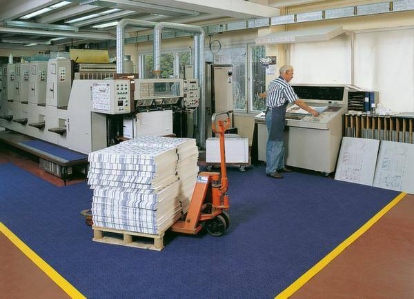 Un homme fait fonctionner des machines d'impression dans une imprimerie. Au premier plan, une pile de papier est posée sur une palette à côté d'un chariot élévateur.