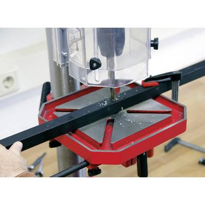 Un morceau de métal est fixé sur un support de perceuse rouge et noir. Un carter de protection entoure la perceuse qui se trouve au-dessus de la pièce.