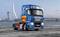 Un camion bleu MAN sans remorque est stationné sur une place urbaine avec un arrière-plan architectural moderne et un pont, sous un ciel nuageux.