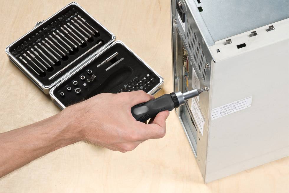 Une main utilise un tournevis pour desserrer une vis à l'arrière d'un boîtier d'ordinateur. Une boîte à outils est posée à côté.