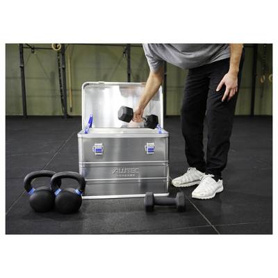 Une personne place un haltère à l'intérieur d'une boîte de rangement métallique dans un environnement de salle de sport, avec des kettlebells et un autre haltère à proximité sur le sol.