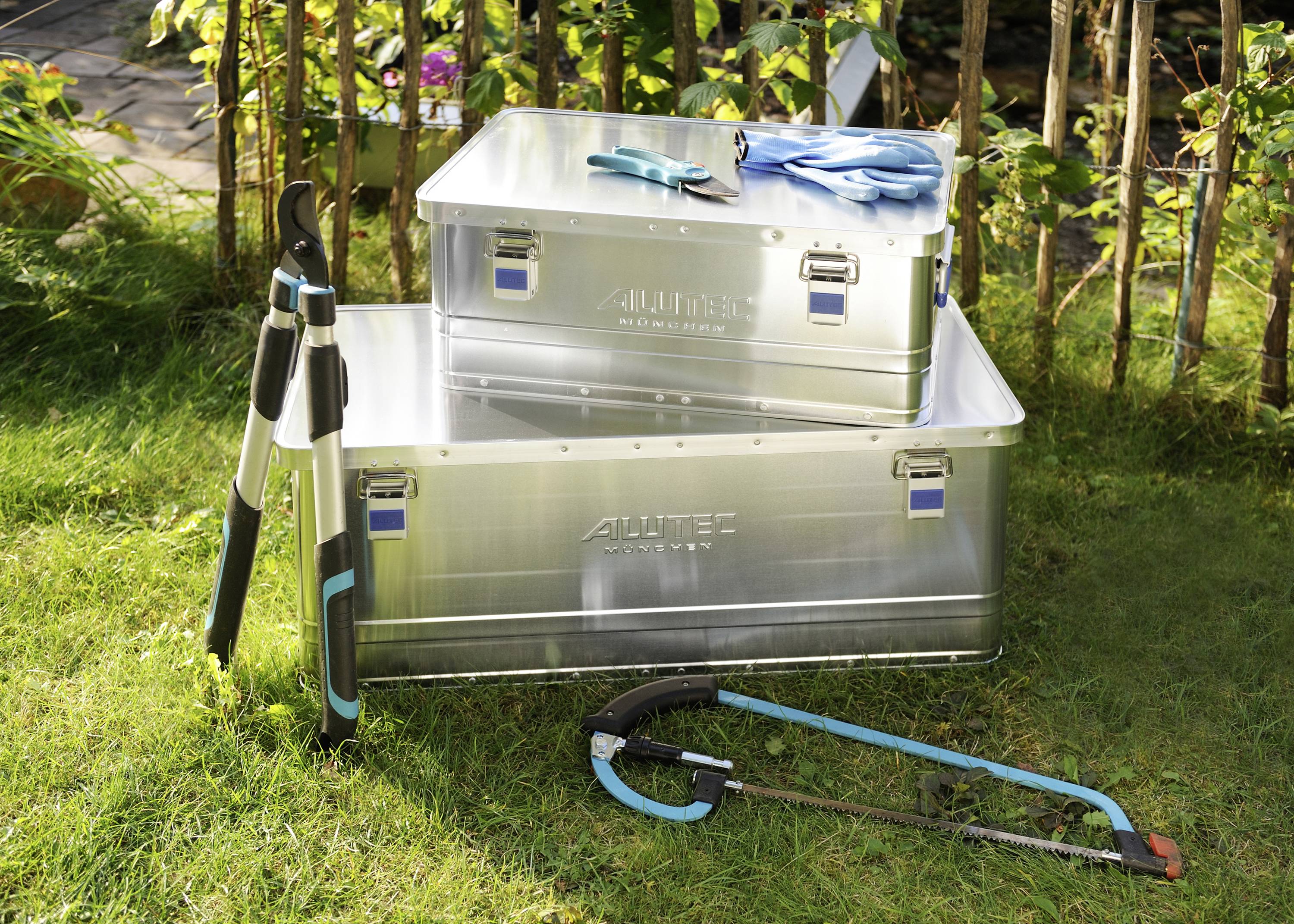 Deux boîtes en aluminium sur une pelouse, entourées d'outils de jardin tels qu'un sécateur et un tuyau d'arrosage, avec une clôture en bois et des plantes en arrière-plan.
