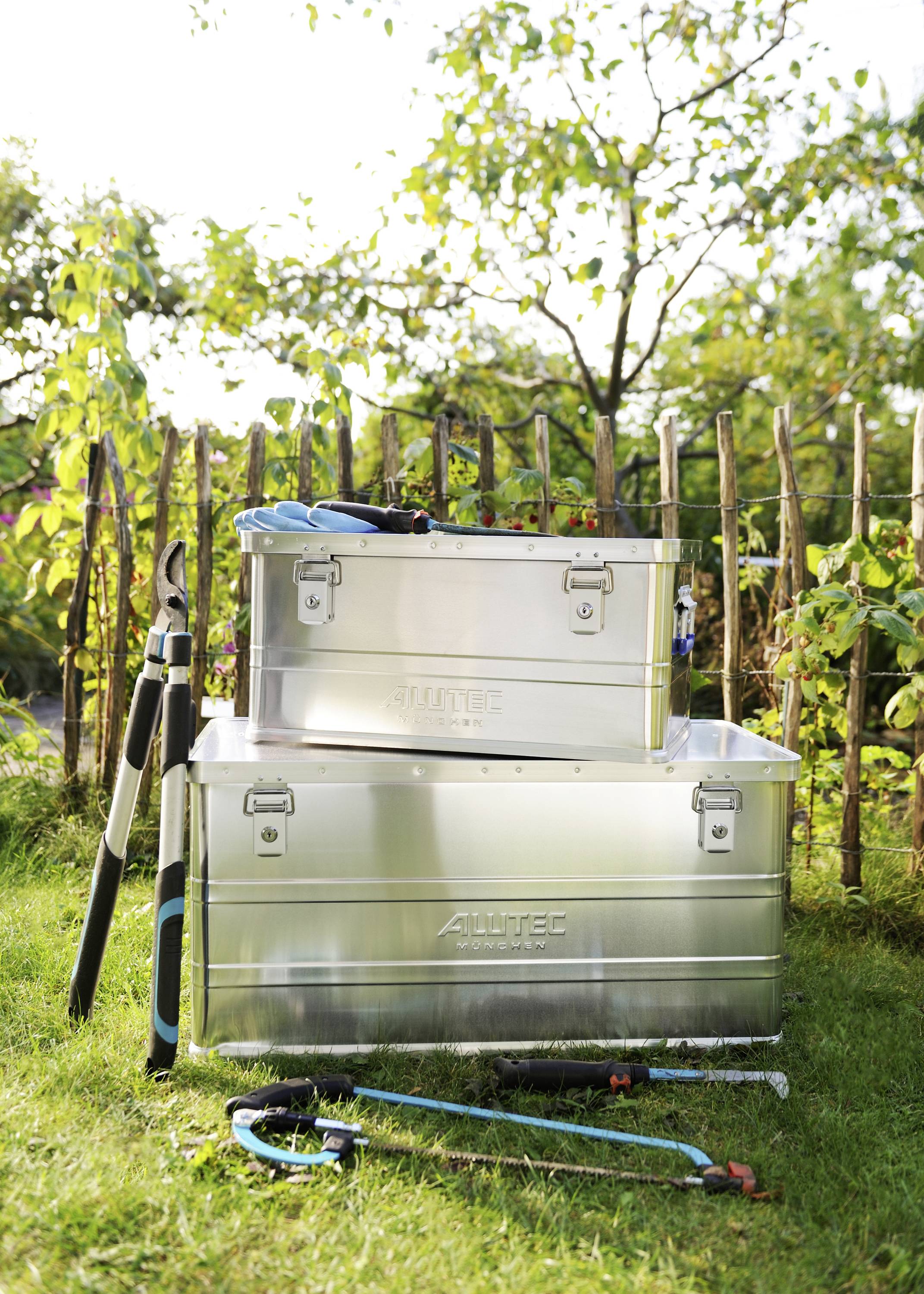 Deux boîtes en métal et des outils de jardin sont posés sur la pelouse devant une clôture en bois. Parfait pour le rangement et l'organisation du jardin.