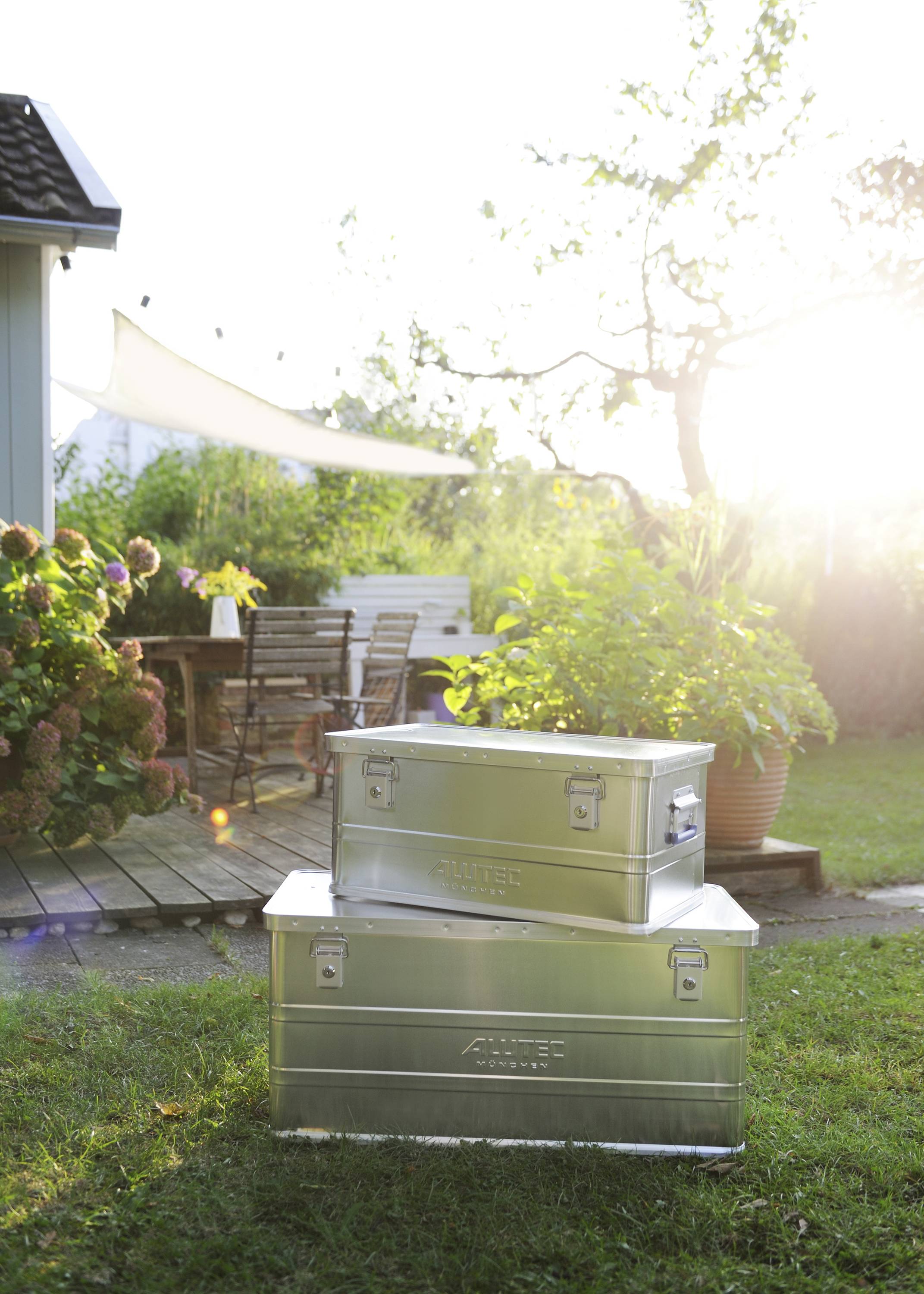 Deux boîtes en métal argenté sont empilées sur de l'herbe verte devant une terrasse avec une table et des chaises en bois sous la lumière du soleil.