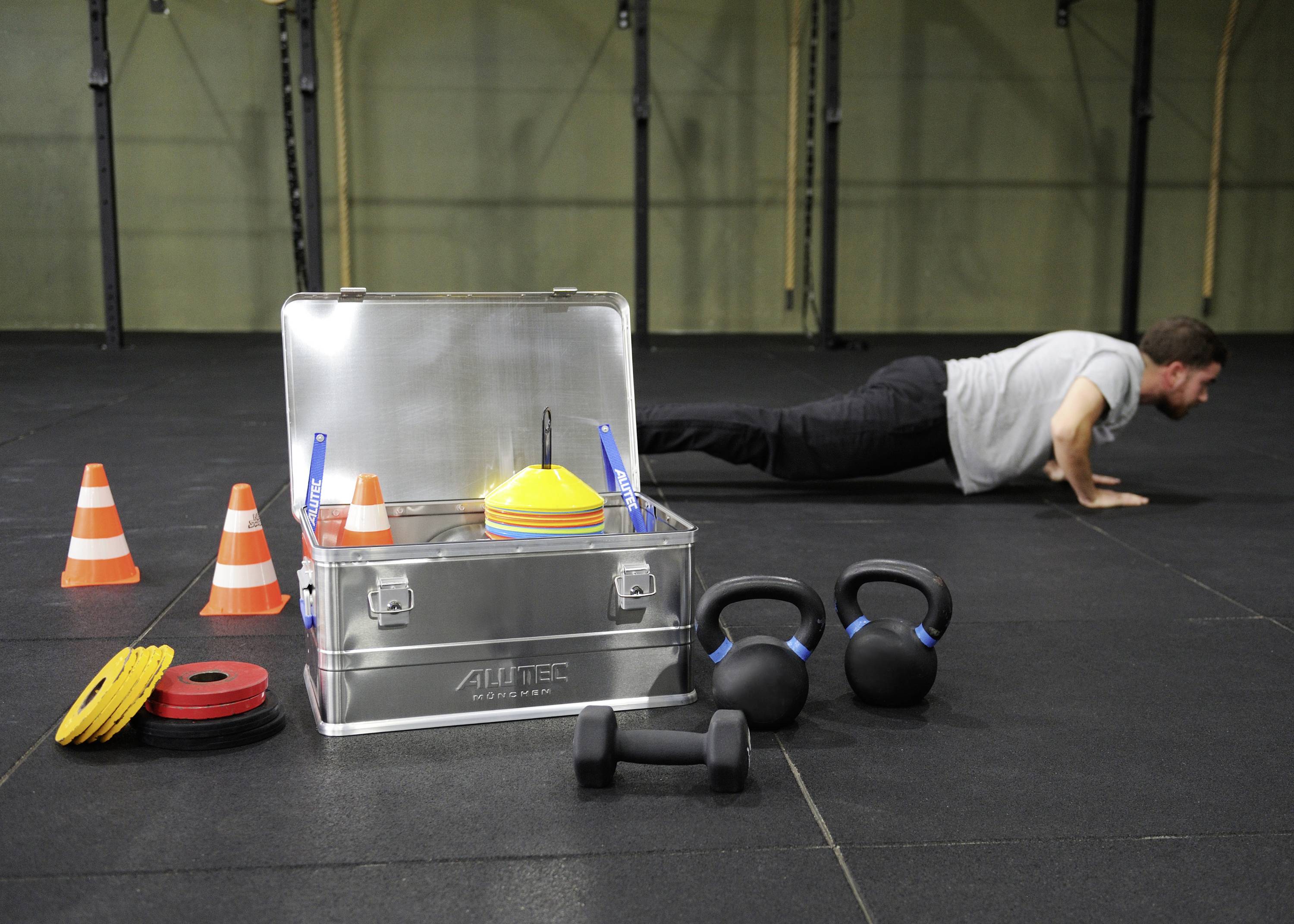 Un homme fait des pompes dans une salle de fitness. À côté de lui se trouvent des kettlebells, des cônes colorés et un coffret métallique ouvert rempli d'équipements sportifs.