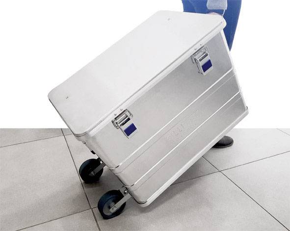 Valise de transport métallique à roulettes inclinée. Elle possède deux fermetures et est posée sur un sol carrelé clair.