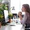Une femme est assise, l'air pensif, à un bureau dans un bureau, avec des plantes et un écran d'ordinateur devant elle.