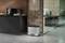 Cuisine avec une machine à café sur un meuble noir ; à côté se trouve une poubelle argentée. Au fond, un bureau vitré.