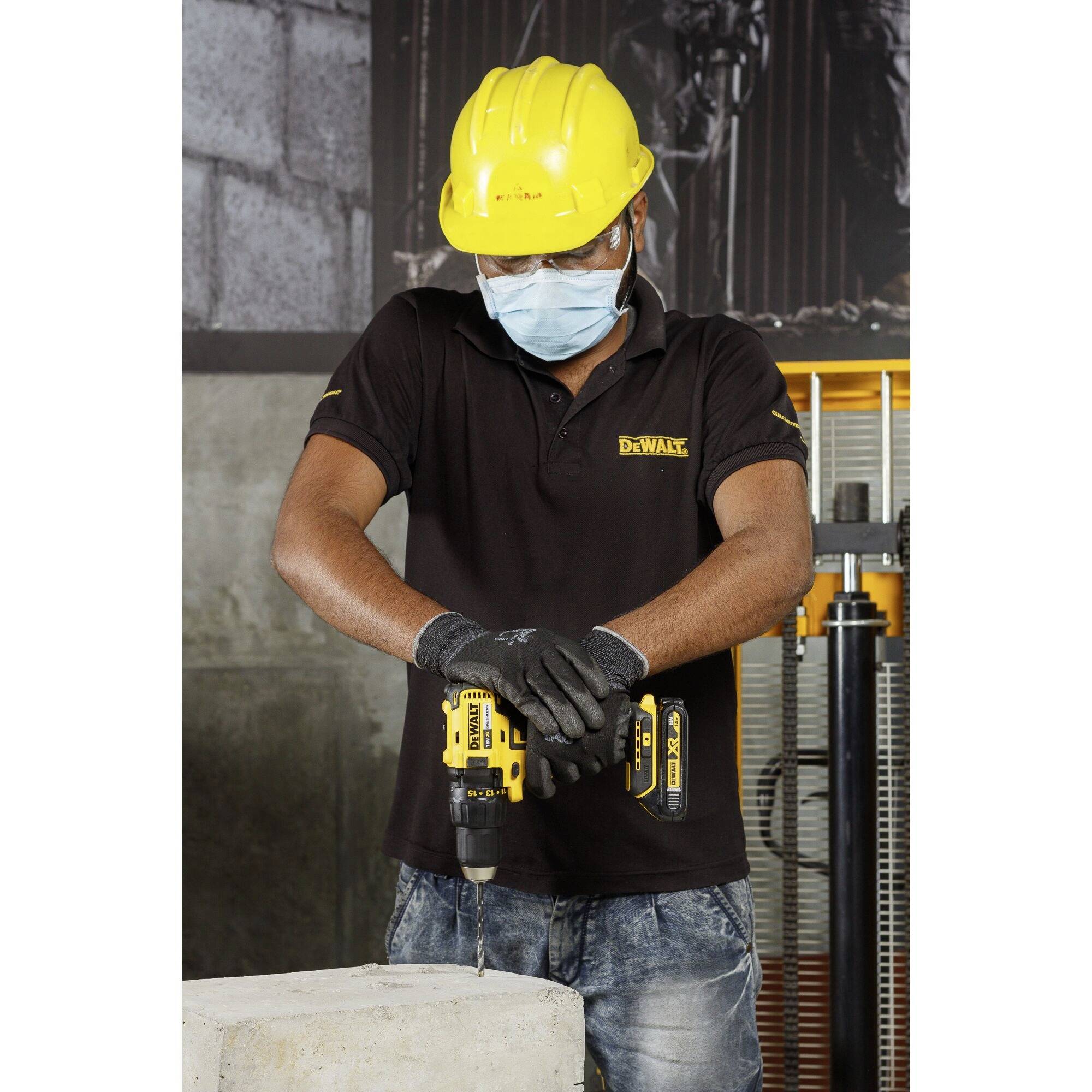 Un ouvrier en tenue de protection fore du béton avec un outil de forage jaune. Il porte un casque, un masque et des gants.