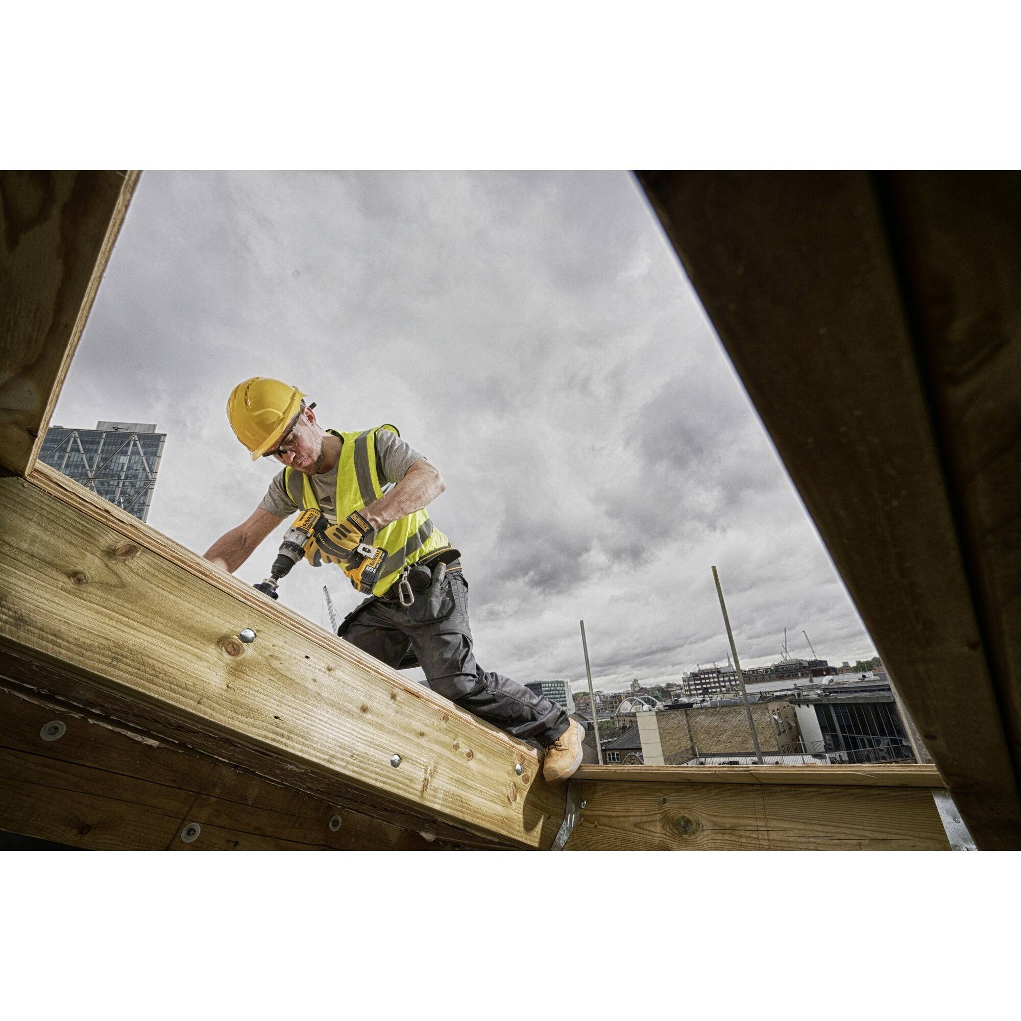 Un ouvrier portant un casque de chantier et un gilet de signalisation fixe des planches de bois à l'aide d'une perceuse sur un chantier sous un ciel nuageux.