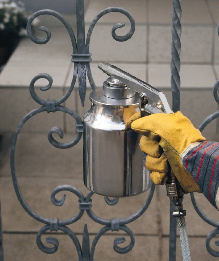 Une personne portant des gants jaunes peint une grille métallique avec un pistolet de pulvérisation argenté.