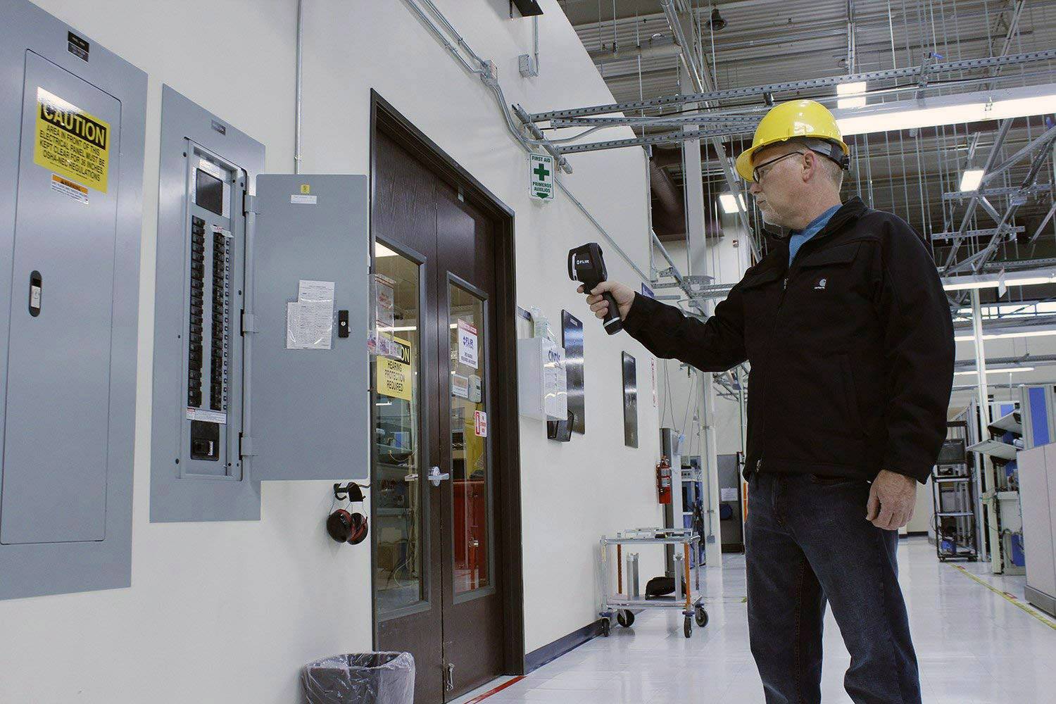 Un homme portant un casque de protection utilise un appareil de mesure infrarouge pour vérifier la température des boîtes de connexion dans un environnement industriel.