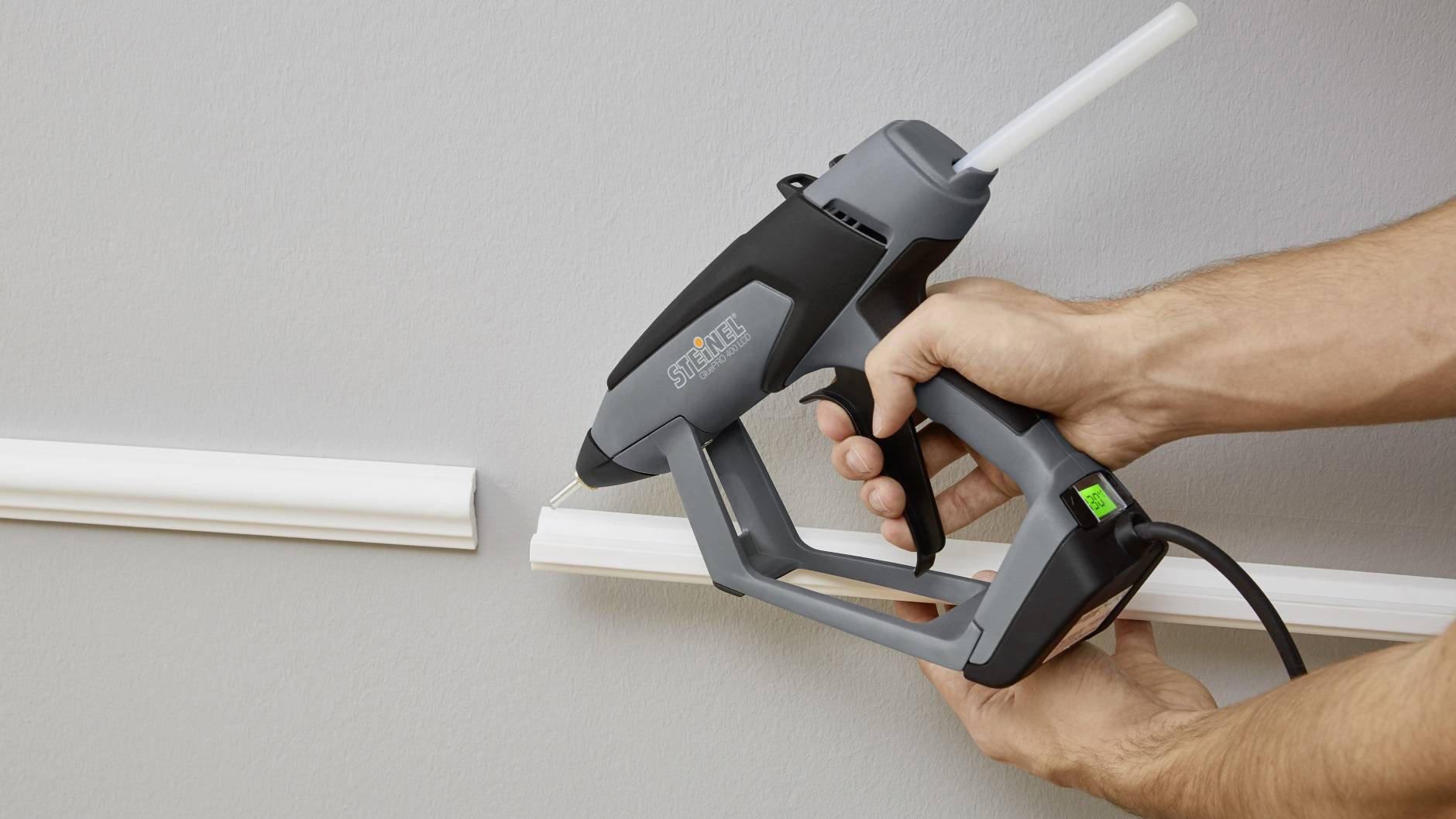 Une personne utilise un pistolet à colle chaude pour fixer une bordure décorative blanche sur un mur gris clair.