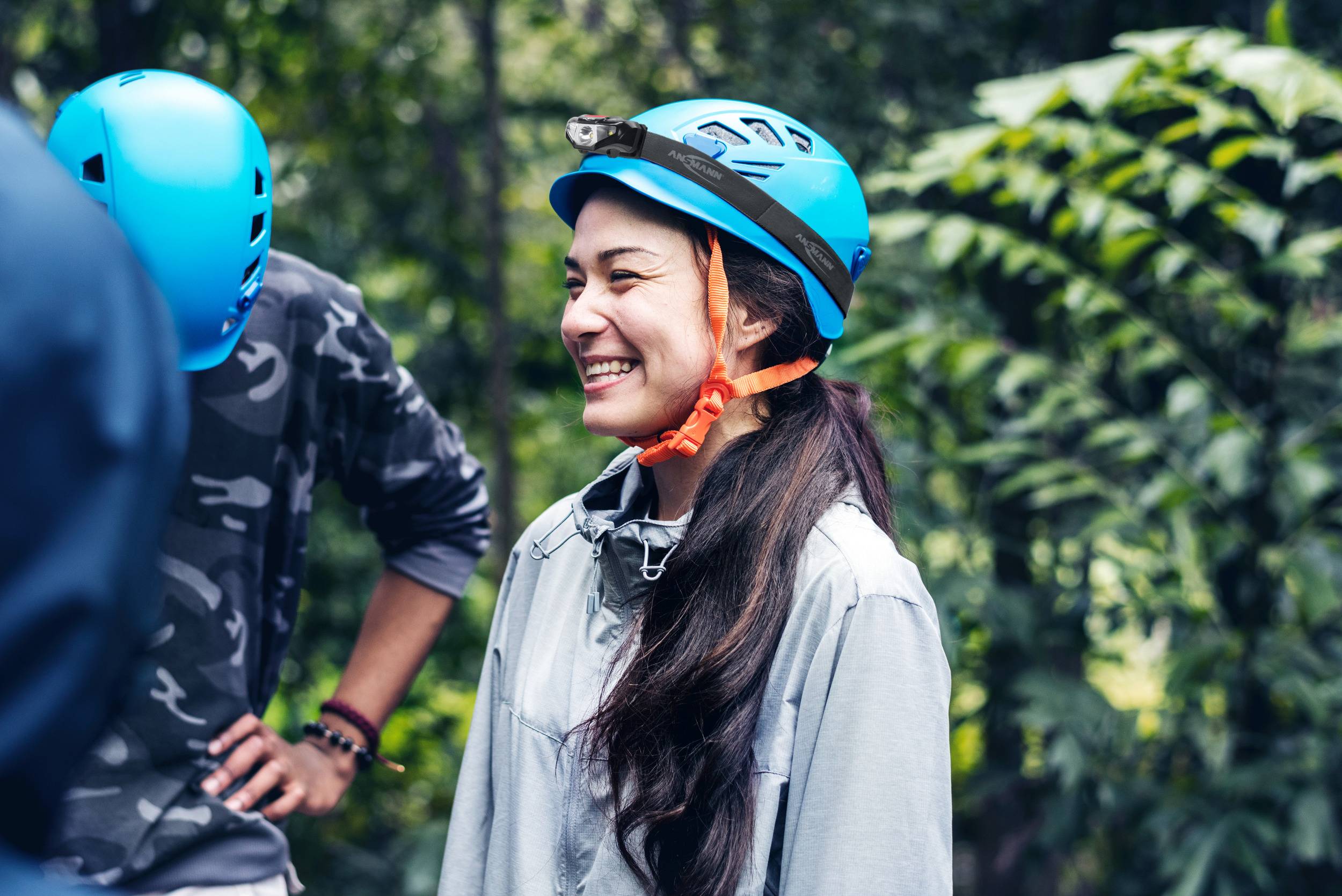 Une personne sourit dans la forêt, portant un casque bleu. Entourée d'autres personnes et d'une végétation luxuriante.