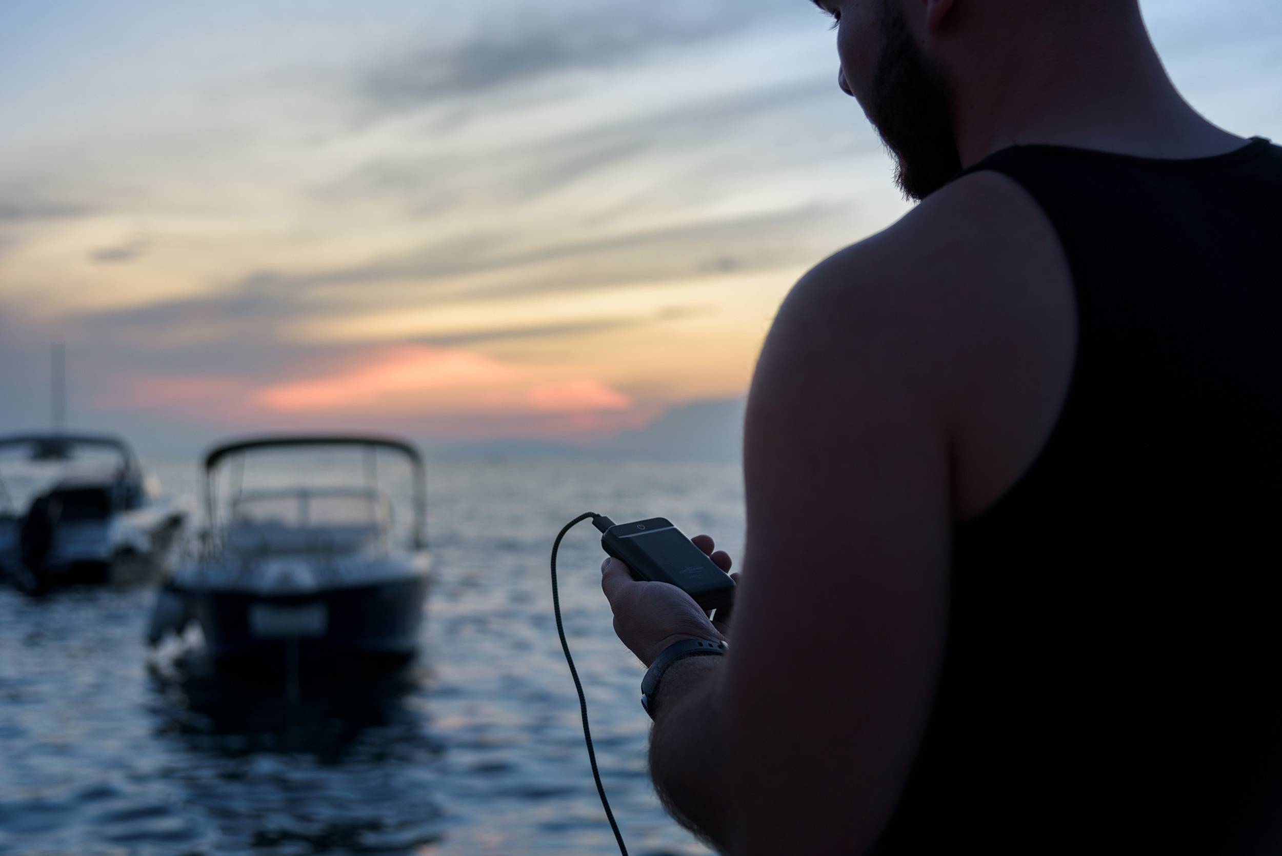 Une personne tient un smartphone à la main et se tient sur la rive avec des bateaux dans l'eau. Le ciel affiche un ciel rouge-orangé aux couleurs chatoyantes du soir.