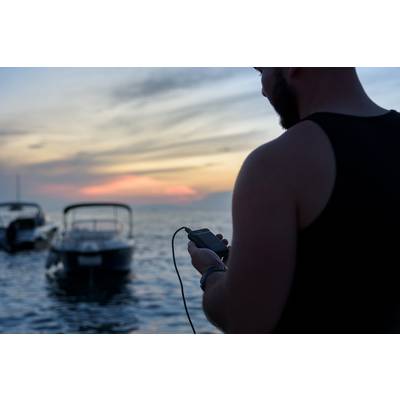 Une personne tient un smartphone à la main et se tient sur la rive avec des bateaux dans l'eau. Le ciel affiche un ciel rouge-orangé aux couleurs chatoyantes du soir.
