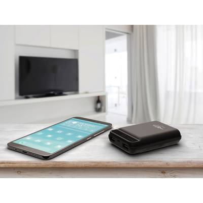 Un smartphone et une batterie externe sont posés sur une table en bois dans un salon moderne. Un téléviseur est visible en arrière-plan.