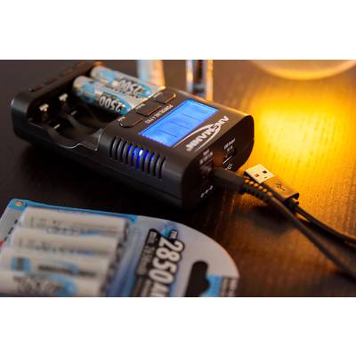 Un chargeur charge des batteries, indiqué par un affichage bleu lumineux. À côté, des batteries de rechange sont posées sur un emballage.