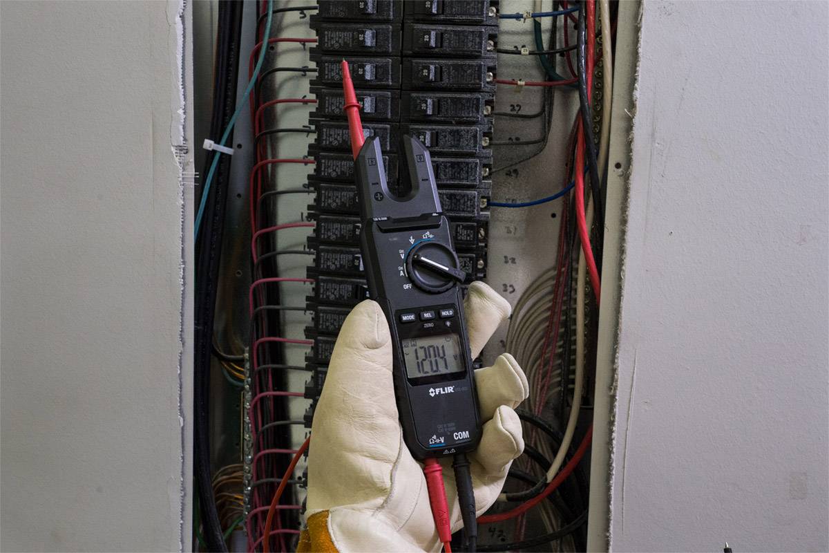 Une personne tient un multimètre mesurant une tension de 120V sur un boîtier de fusibles ouvert. Plusieurs câbles sont visibles.
