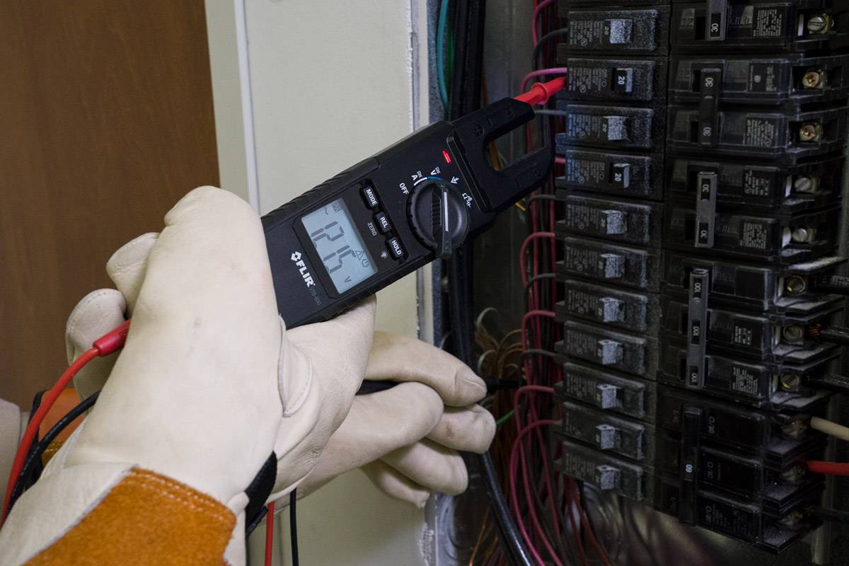 Une personne portant des gants de protection effectue un test de tension avec un appareil de mesure électrique dans le tableau électrique.