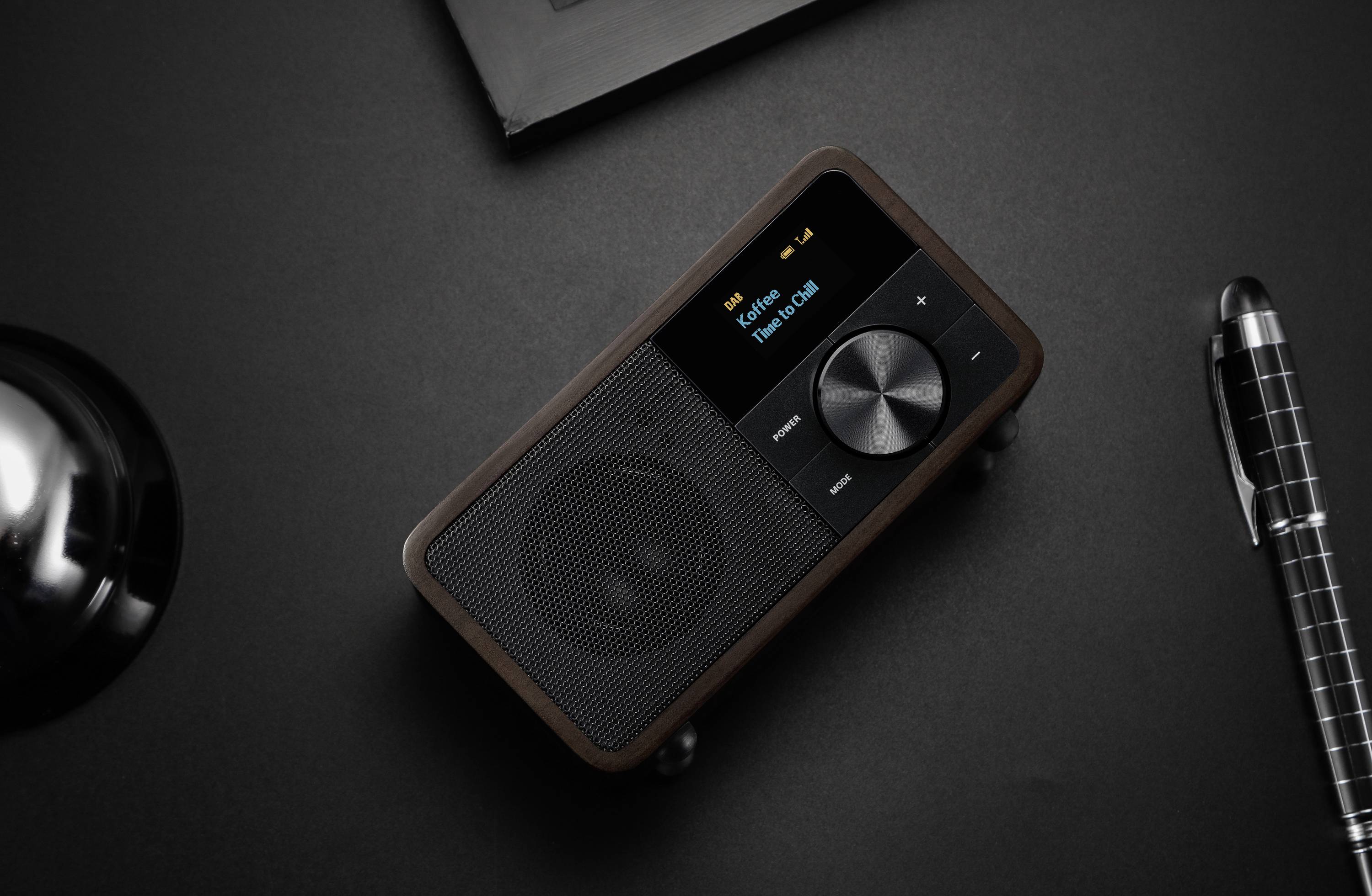 Un haut-parleur portable avec un cadre en bois, un réglage de volume et un écran affichant le titre d'une chanson, est posé sur une table.