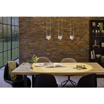 Salle à manger avec une table en bois, cinq chaises et des livres sur la table. Trois lampes sont suspendues au-dessus de la table. De grandes fenêtres donnent sur un vue de jardin.